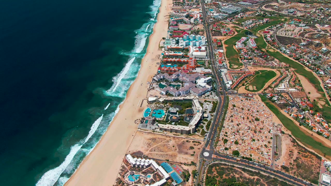 San Jose Del Cabo aerial view showing the shoreline running vertically through frame, ocean surf on the left, resort complexes beside the beach, a road and roundabout, and golf greens to the right with no visible people.