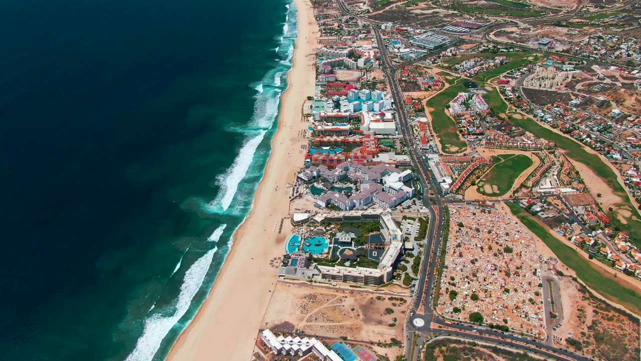 Aerial view over San Jose Del Cabo with dark blue ocean at left, white surf reaching a sandy beach, resort-style buildings, roadways, and green grounds to the right, with no visible people.
