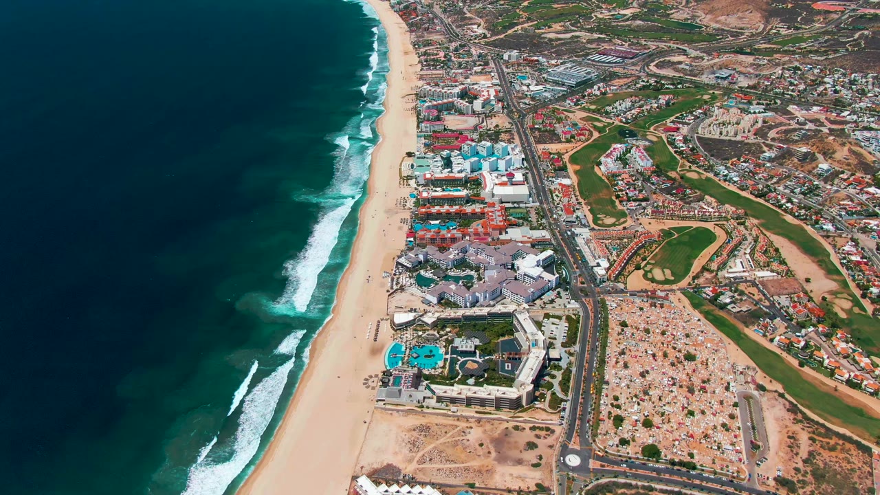 Aerial frame of San Jose Del Cabo where white wave lines roll toward the sand beside clustered coastal buildings, turquoise pools, a road, and dry hillside construction, with no visible people.