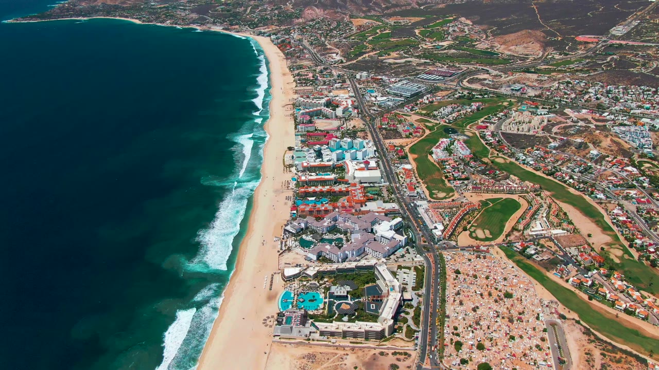 Opening aerial frame over San Jose Del Cabo, Baja California Sur, with dark ocean at left, surf on the sand, a coastal highway, resort buildings, green golf areas, and no visible people.