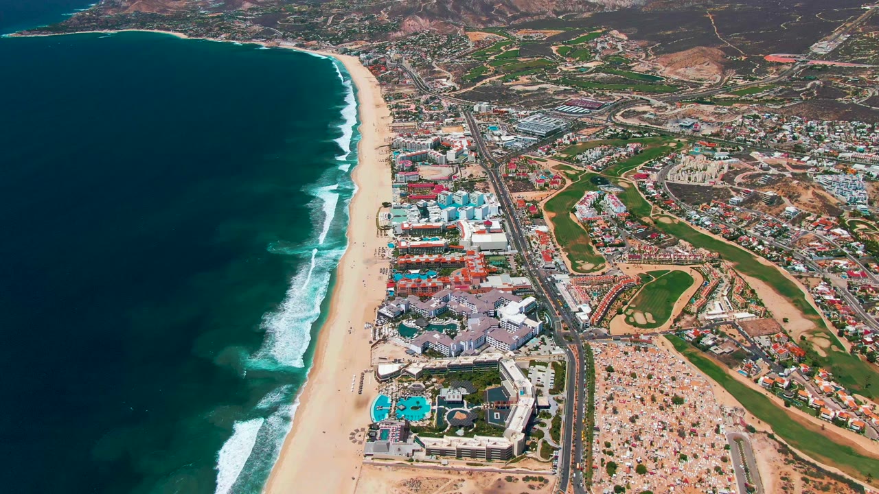 Aerial frame in San Jose Del Cabo showing the beach and surf line beside the coastal road, with small vehicles, hotel-style buildings, green fairways, inland streets, and no visible people.
