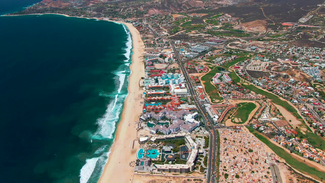 Wide aerial view of blue water, beach, buildings, highway traffic, and golf course.