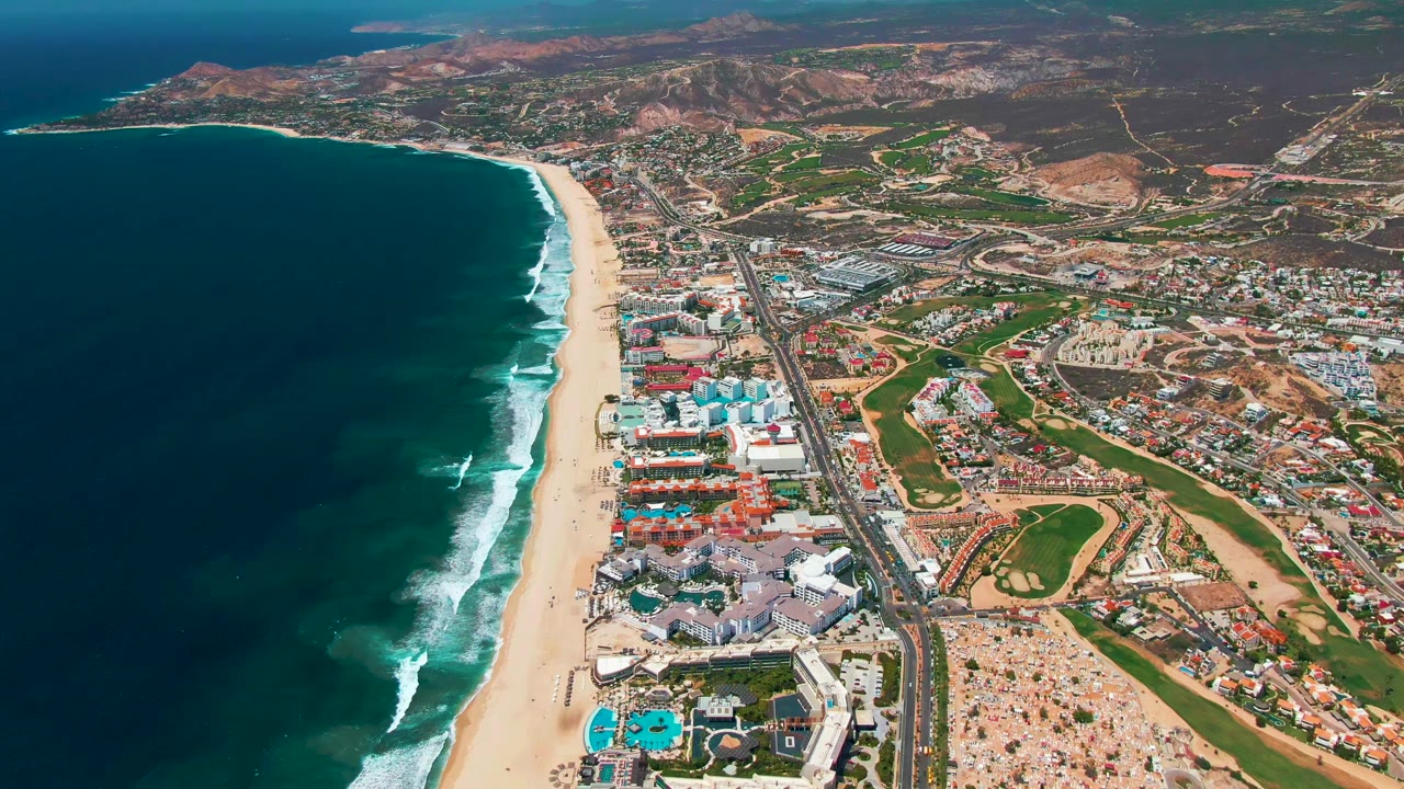The coastline in San Jose Del Cabo curves across the frame, with dark ocean water, staggered white surf, a wide sandy beach, large seaside buildings, roads, inland lots, reddish hills, and no visible people.