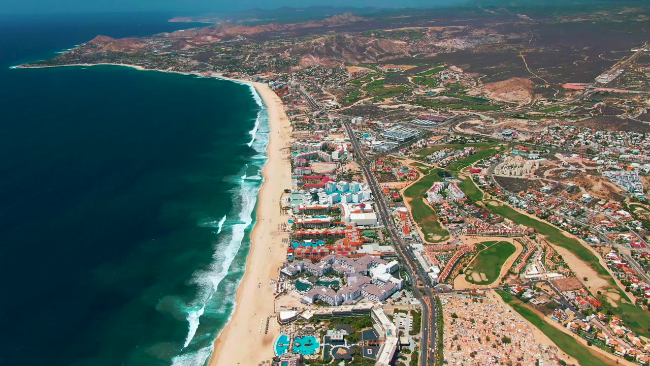 Drone-style coastal view with ocean, surf, beach, buildings, road, and hills.