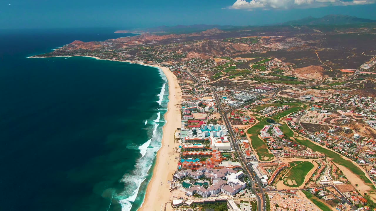 At 0:00, a high aerial view over San Jose Del Cabo shows dark blue ocean, a long sandy beach with white surf, coastal buildings, a road, green landscaped areas, dry hills, and no visible people.