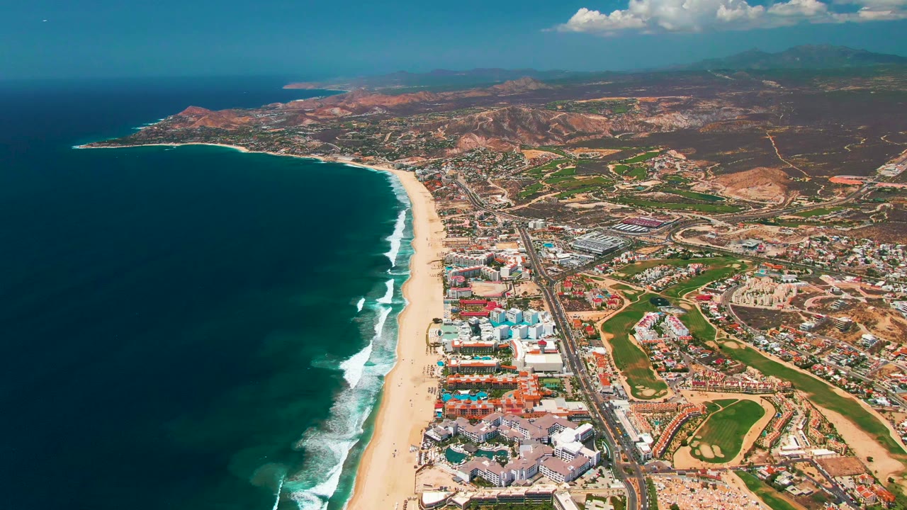 At 0:01, the San Jose Del Cabo shoreline remains framed from above, with surf bands advancing along the beach, small vehicles on the coastal road, clustered buildings, inland hills, and no visible people.