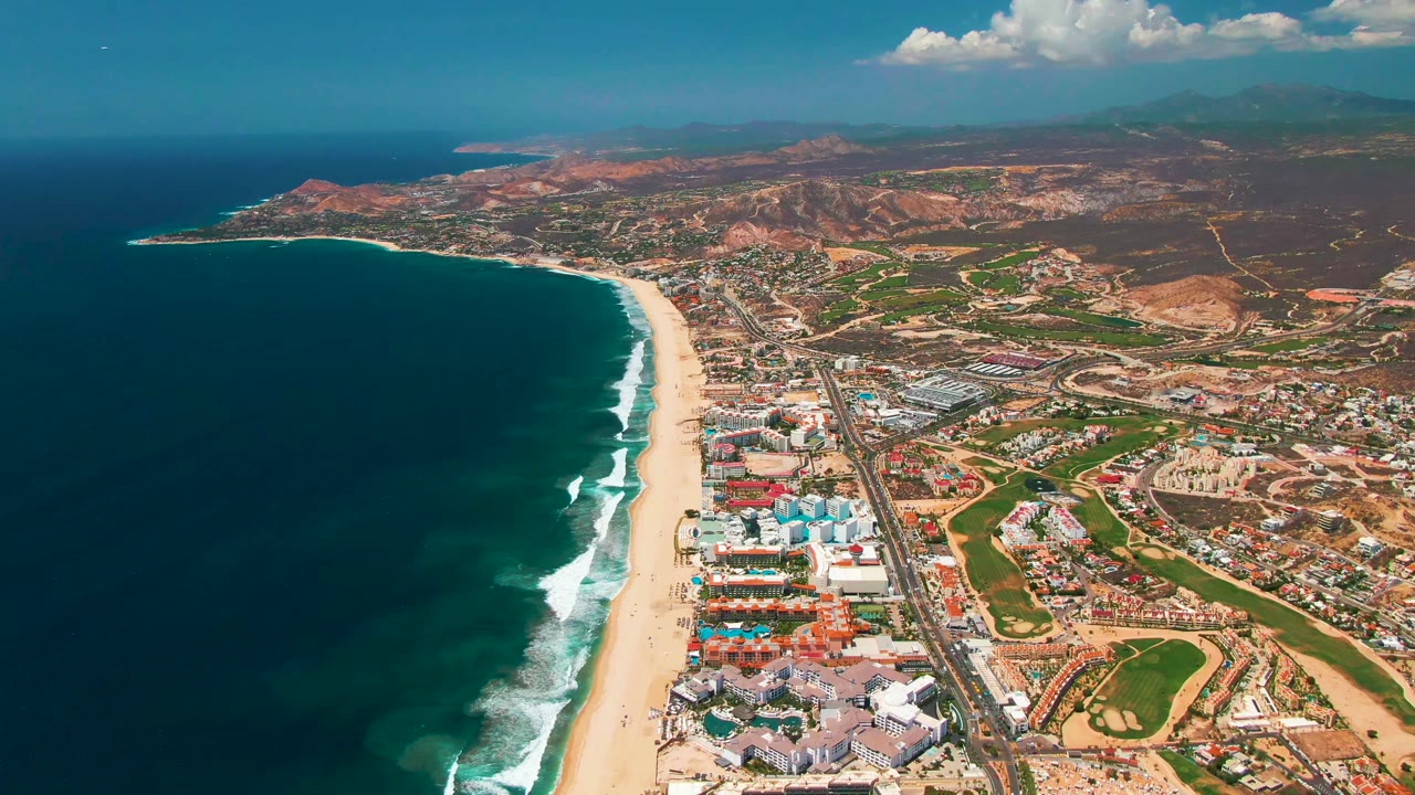 At 0:02, the aerial angle looks along the San Jose Del Cabo coast with the beach curving into the distance, dark ocean at left, town buildings and road at right, inland hills beyond, and no visible people.