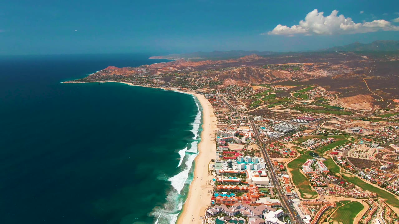 A wide aerial frame over San Jose Del Cabo shows dark blue ocean at left, a pale sandy beach, beachfront buildings, roads, green fields, inland hills, scattered clouds, and no visible people.