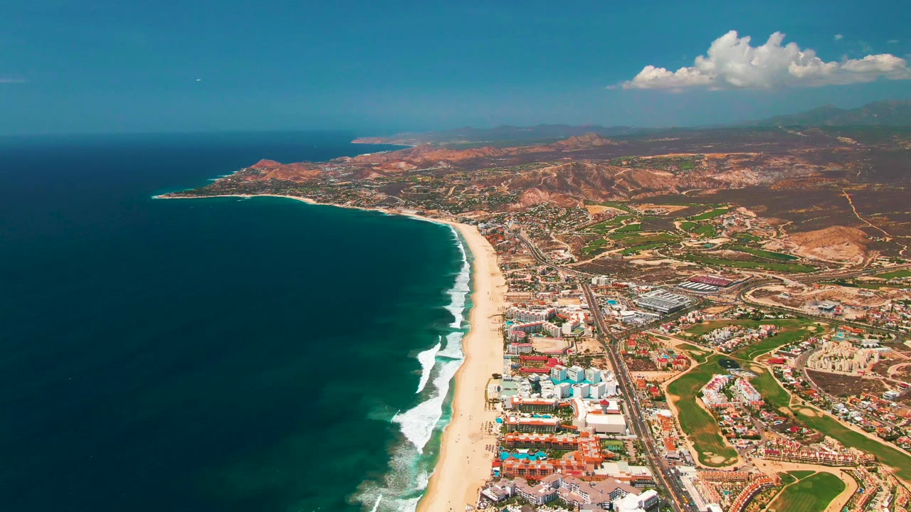 At 0:01, the San Jose Del Cabo shoreline curves through the frame with deep blue water, white wave lines, sandy beach, low-rise development, roads, green patches, arid hills, and no visible people.