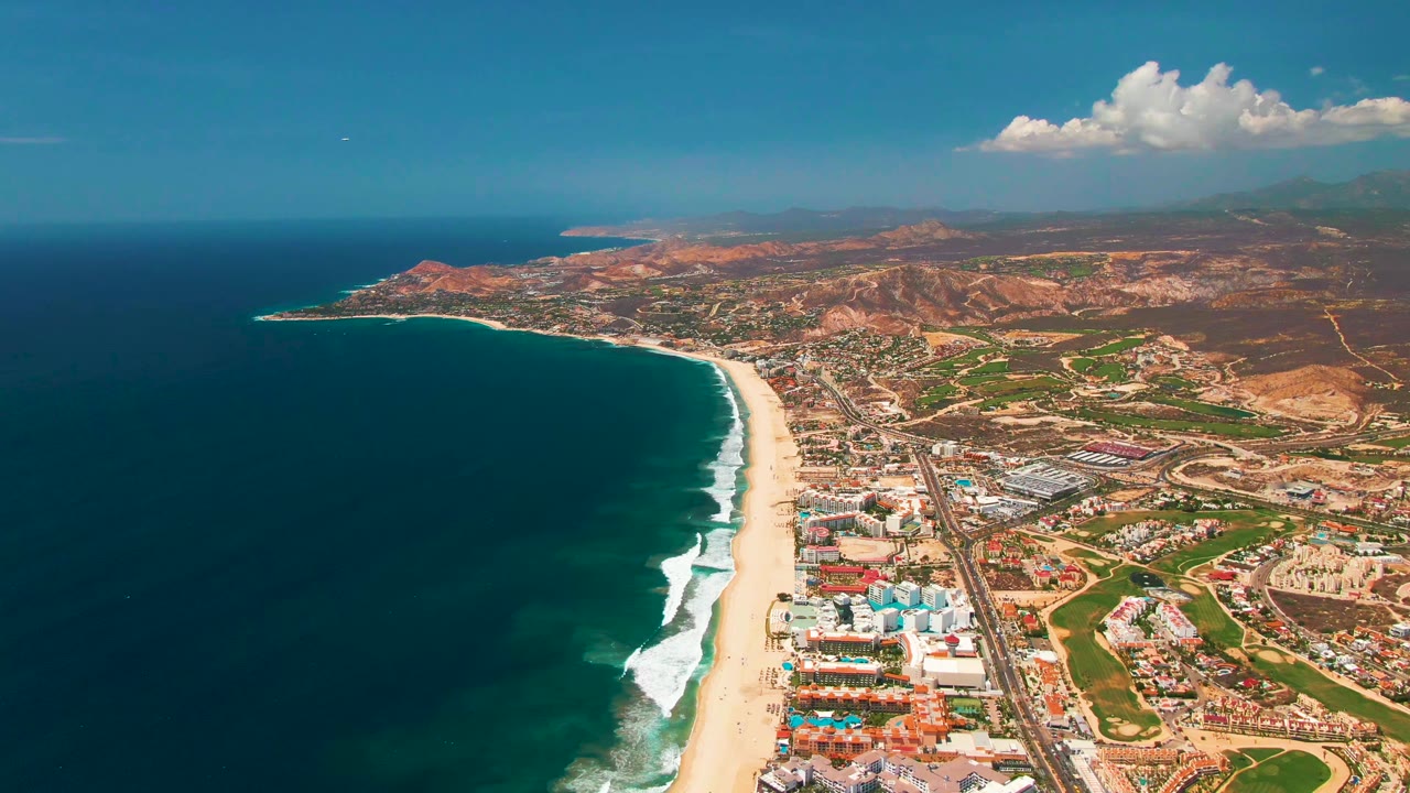 High aerial coastline with beach, ocean, buildings, roads, golf-like green areas, and hills.