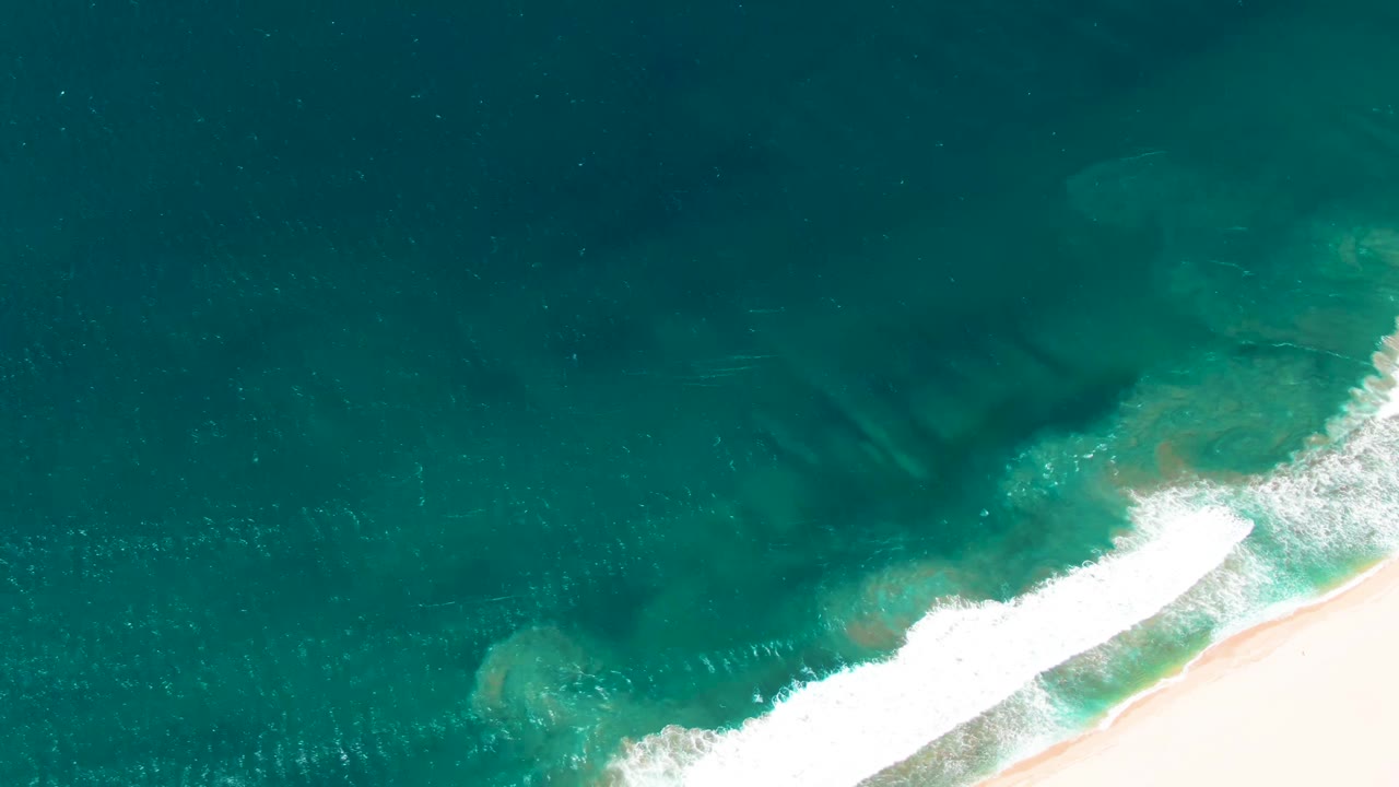 Overhead view in San Jose del Cabo showing dark blue-green water, turquoise shallows, white surf, pale sand, and no visible people at the start of the clip.