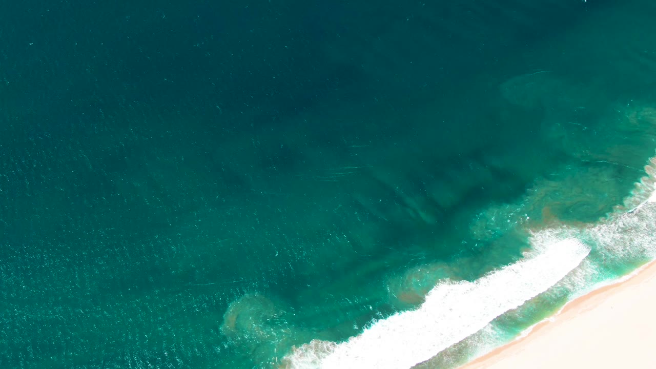 Aerial surf line with white foam moving toward pale sand.
