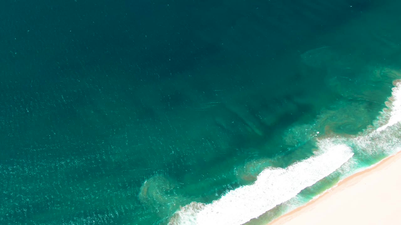 Overhead San Jose del Cabo coastline with a thicker white wave band crossing the lower right shoreline, darker water offshore, and no visible people.