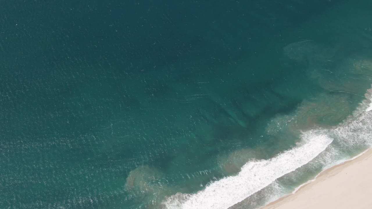 Aerial view in San Jose del Cabo shows teal ocean water, a diagonal band of white surf, pale sand at the lower right, and no visible people.
