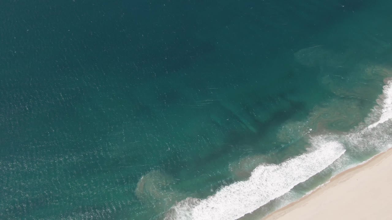 Overhead beach view in San Jose del Cabo with a curved white breaker crossing turquoise water toward pale sand, with no visible people in the frame.