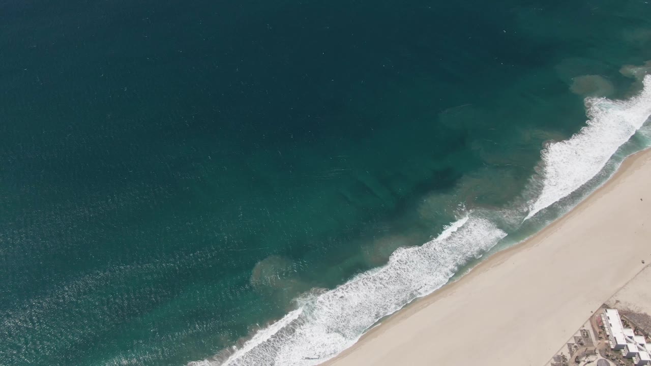 Aerial frame over San Jose del Cabo showing turquoise ocean and white surf reaching a diagonal sandy shoreline, with no visible people.