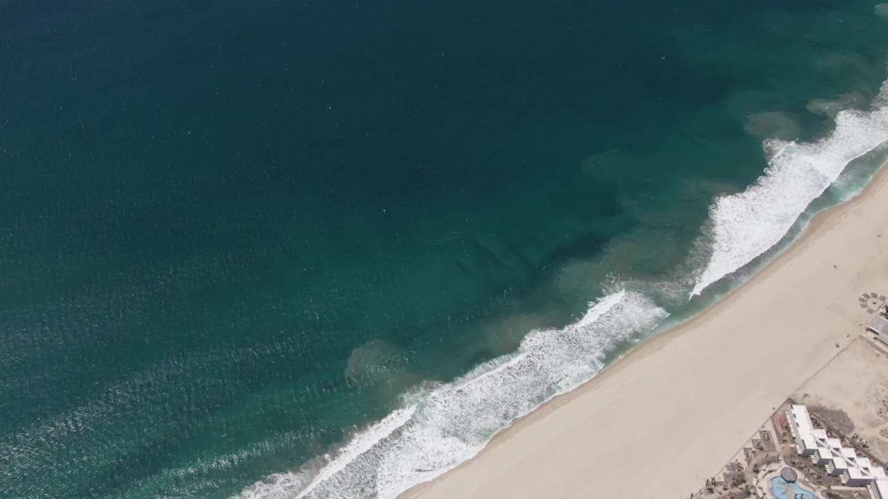 Overhead coastline with white waves rolling toward pale sand.