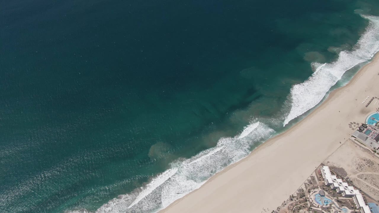Aerial frame in San Jose del Cabo with broad turquoise water, breaking waves, sandy beach, and poolside coastal structures near the right edge, with no visible people.