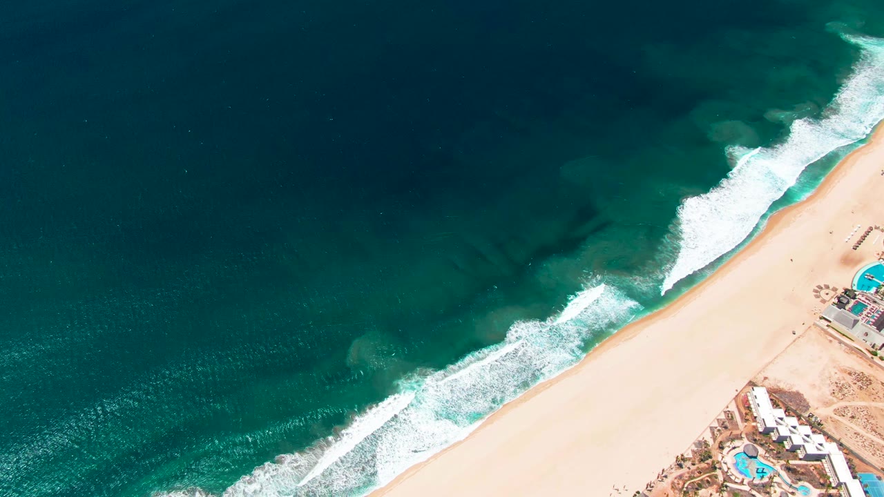 Aerial view of waves breaking along a sandy beach beside resort pools.