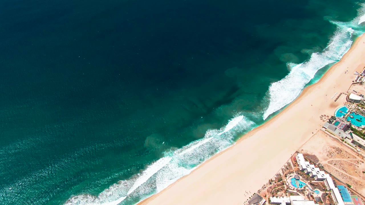 Aerial San Jose del Cabo shoreline frame with a broad dark ocean area, diagonal white breakwater foam, pale sand, and blue resort pools, with no visible people.