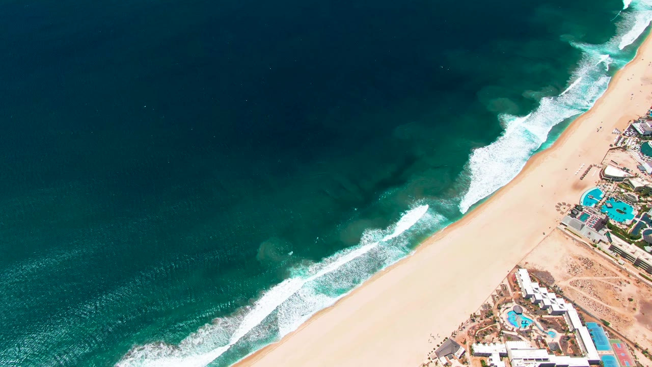 Overhead San Jose del Cabo beach view with foamy surf bands angled across the frame, resort pools clustered on the right, and no visible people on the sand or in the water.