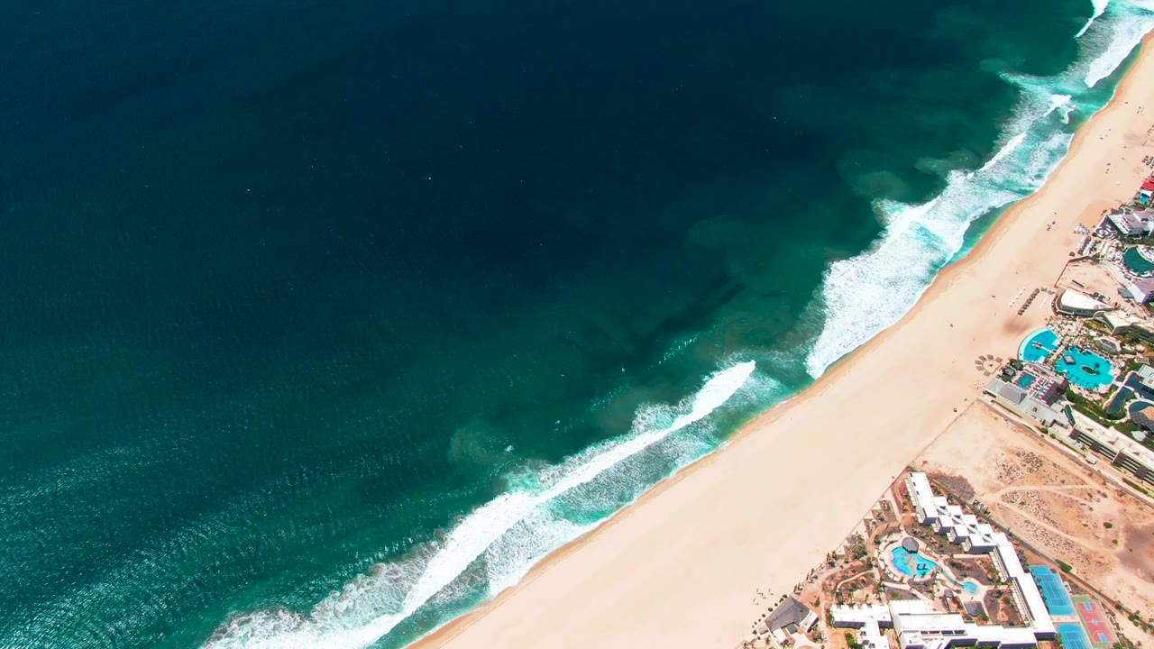 San Jose del Cabo aerial frame showing ocean occupying most of the image, a clean diagonal strip of sand, resort rooftops and pools at right, and no visible people.