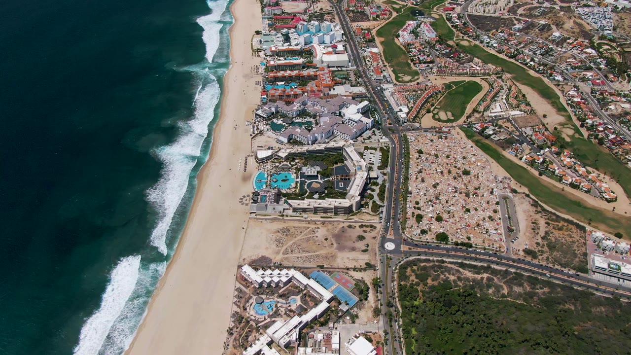 Aerial frame from San Jose del Cabo showing dark ocean surf beside a wide beach, resort buildings and pools near the shore, a central road, parking areas, and golf course greens, with no visible people.
