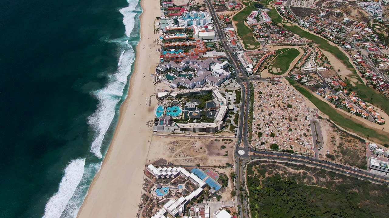 Overhead coastline with waves, sandy beach, resort complex, road, parking, and golf course.