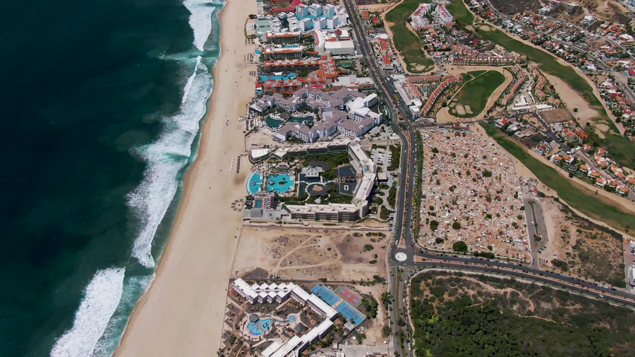 High aerial frame over San Jose del Cabo with the surf line shifted along the left edge, a broad empty-looking beach, resort pools and low buildings below center, a main road, parking area, and golf course features on the right, with no visible people.