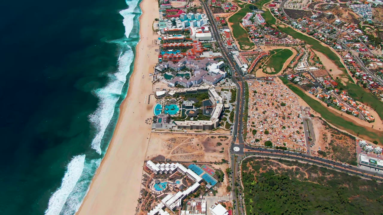 Aerial frame over San Jose Del Cabo, Mexico, showing dark teal surf along a wide sand beach, resort pools near the shore, a parallel road, and a golf course to the right with no visible people.
