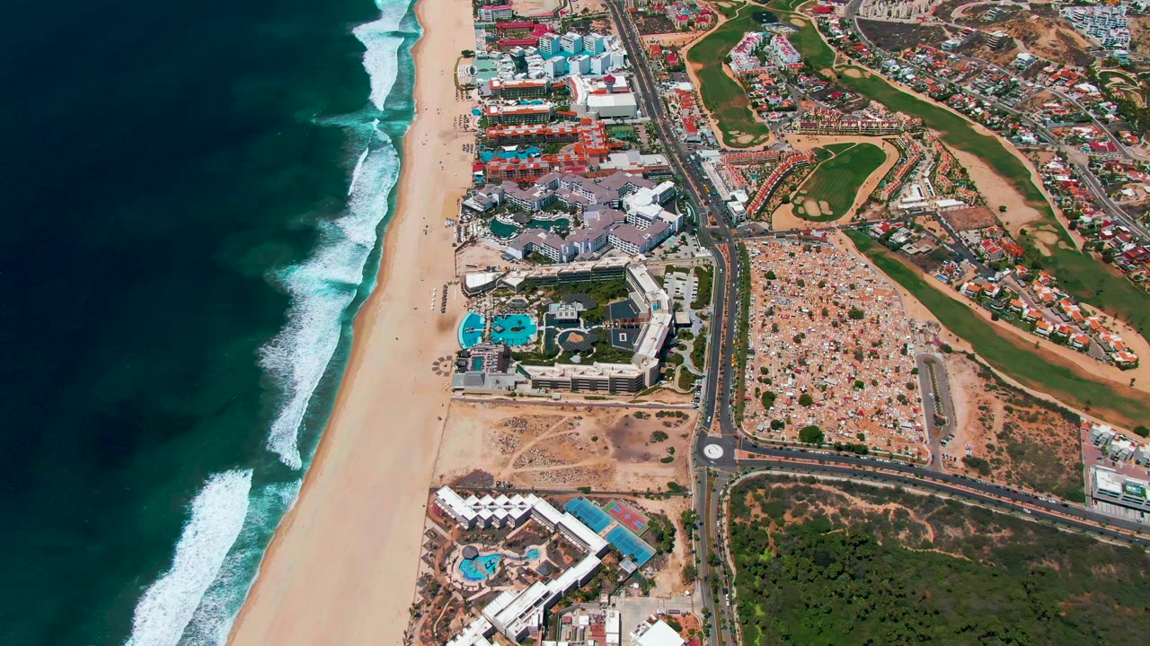 Aerial coastline with surf, beach, resort buildings, road, and green fairways.