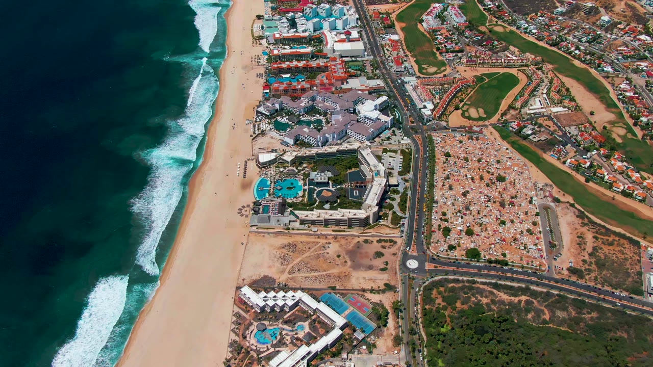 San Jose Del Cabo aerial frame showing ocean and whitecaps along the left side, a long sandy beach, clustered resort buildings and pools, a main road, dense low-rise development, and golf greens on the right with no visible people.