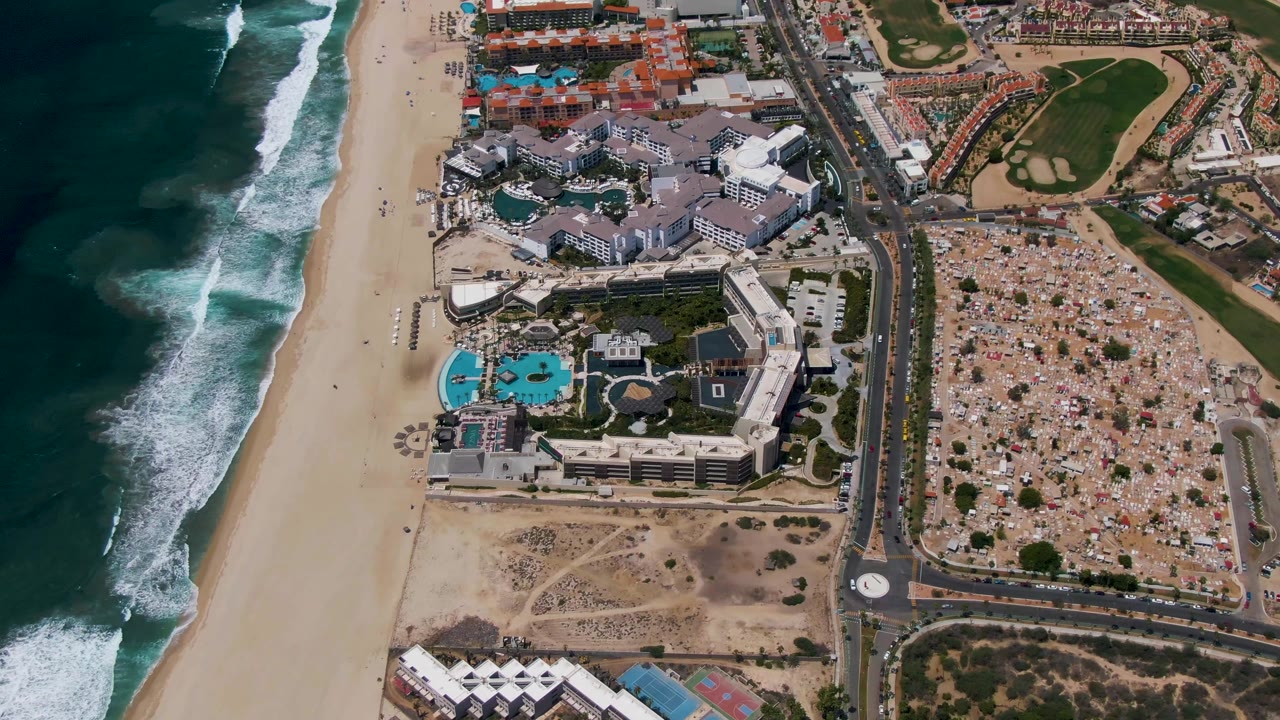 Aerial view of ocean surf, sandy beach, resort buildings, road, roundabout, and golf course area.