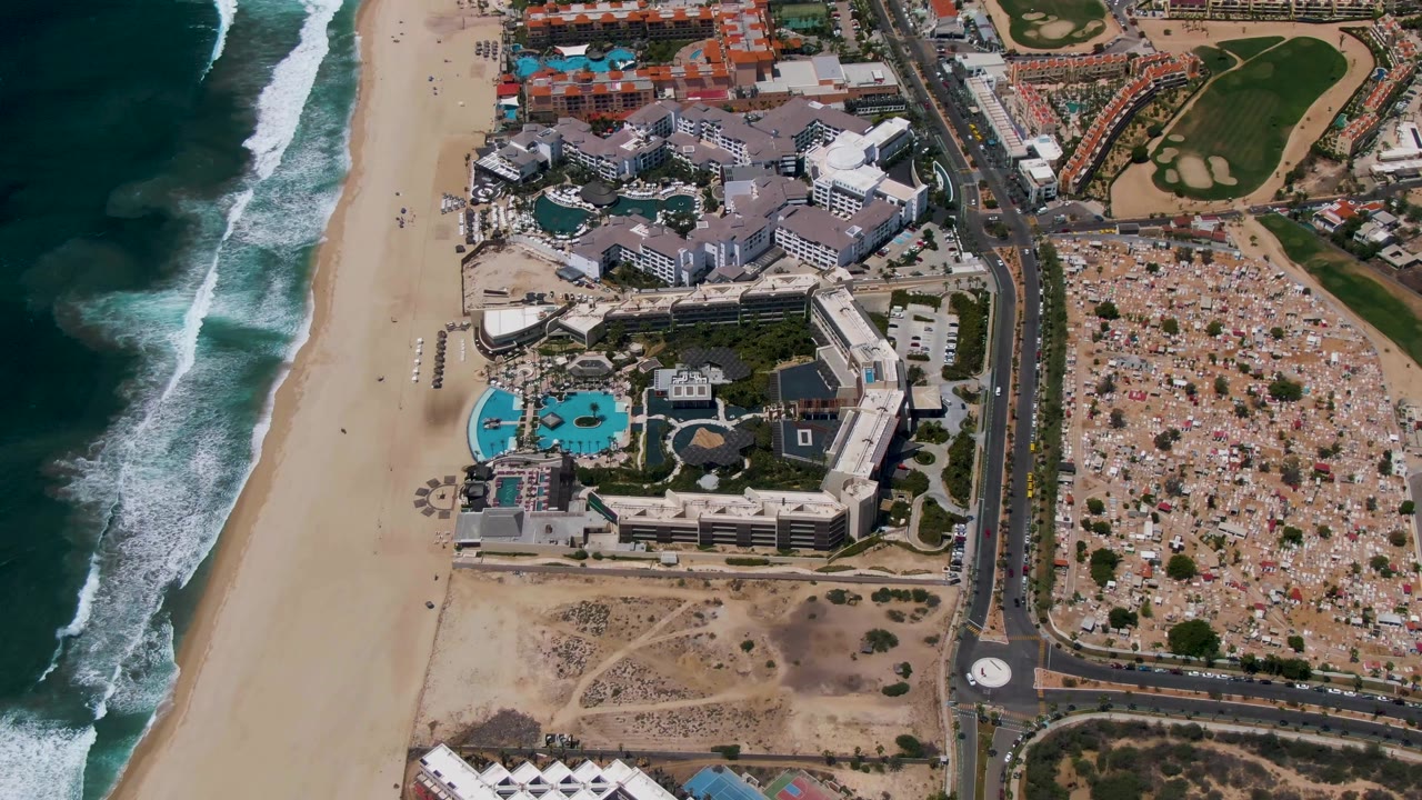 Aerial view in San Jose del Cabo with the surf line shifting along the beach, resort pools near the center, a straight road and roundabout to the right, and green golf course sections beyond, with no visible people.