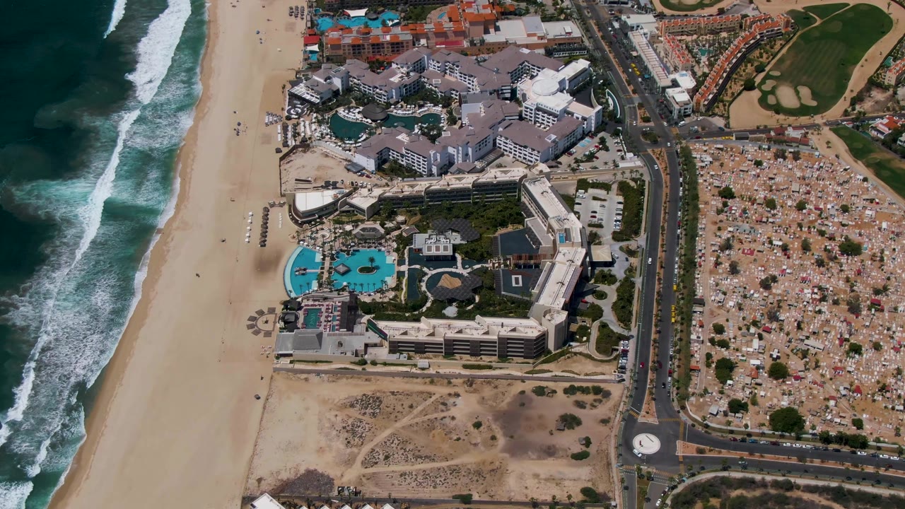 High aerial frame of San Jose del Cabo with ocean waves on the left, the sandy beach and resort complex through the middle, and the roundabout, parking density, and golf course area on the right, with no visible people.