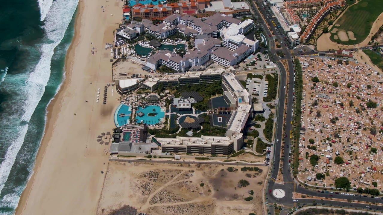 Aerial frame over San Jose Del Cabo showing the beach and waves on the left edge, resort complex and pools near center, and road traffic beside golf greens and tan development, with no visible people.