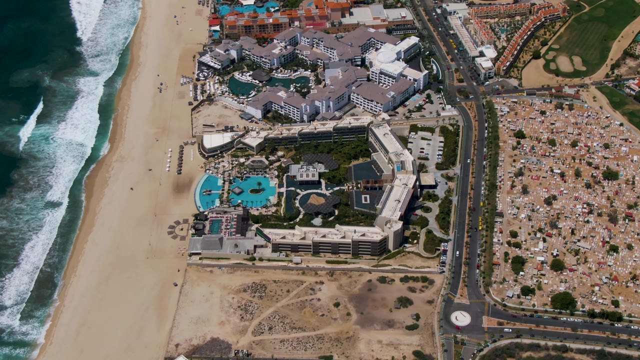 Drone view of beach resort area with ocean surf, pools, road, and construction-like land.