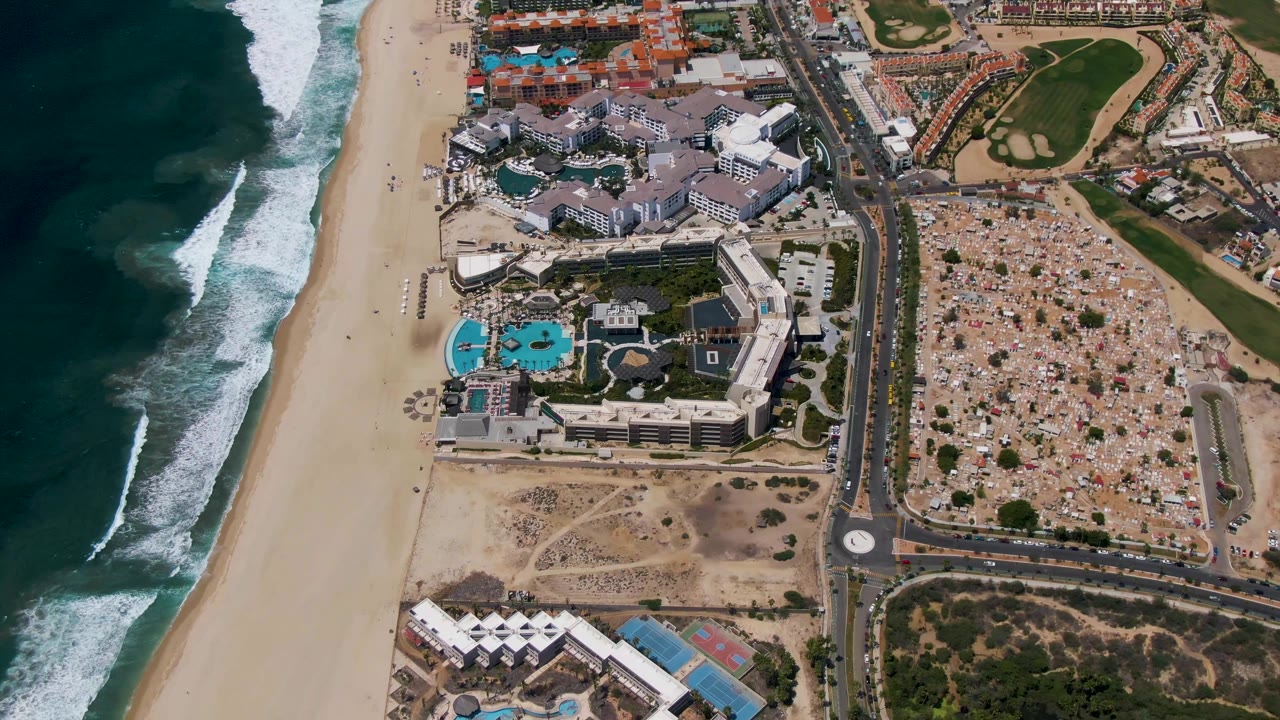 Aerial frame over San Jose Del Cabo showing dark ocean surf at left, a wide sandy beach, resort pools and buildings, roads, parking areas, and no visible people.