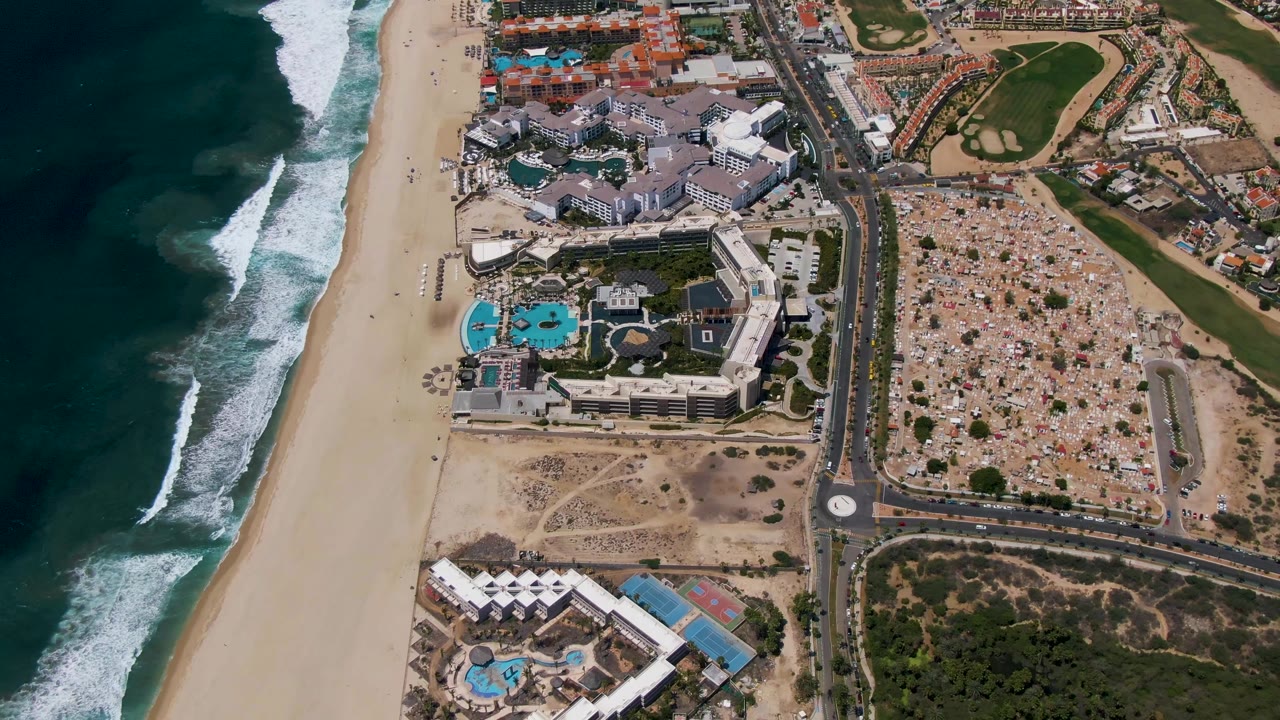 Overhead coastline with surf, resort pools, road, cars, and adjacent developed lots.