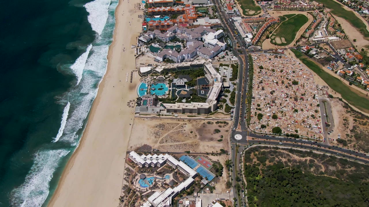 Aerial frame over San Jose Del Cabo showing more open water at left, diagonal white wave breaks, a broad beach, clustered resort roofs, pool courtyards, and no visible people.