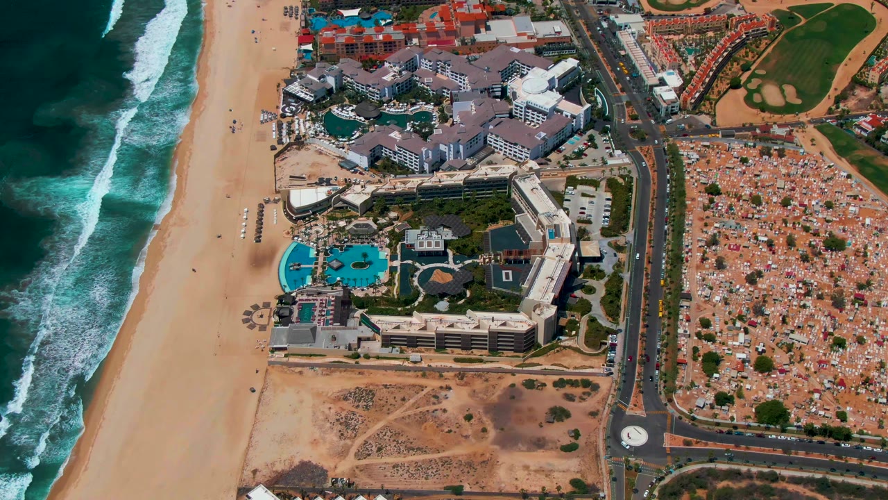 Overhead view in San Jose del Cabo, Mexico, showing waves along a wide beach, resort buildings with blue pools, a road roundabout, parking area, and greener golf course grounds, with no visible people.