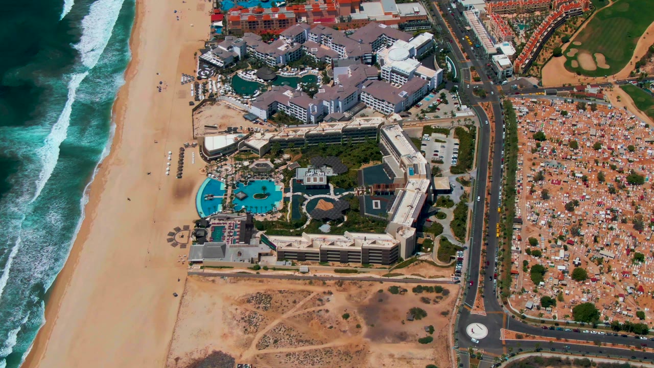 Overhead resort complex beside surf with blue pools, beach, road, roundabout, parking, and brown course area.