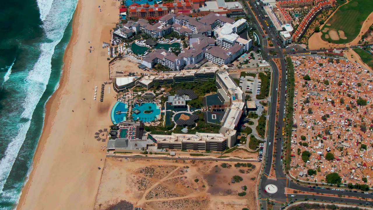Aerial view of beach, ocean waves, resort buildings, pools, roads, and inland lots.