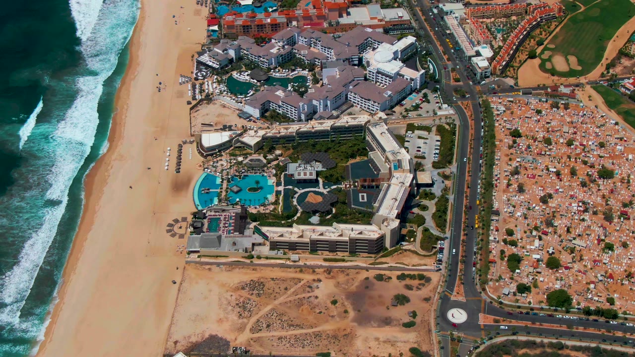 Second aerial frame in San Jose del Cabo with white surf at left, a broad sand beach, central hotel and pool structures, and dense inland development beyond the road, with no visible people.