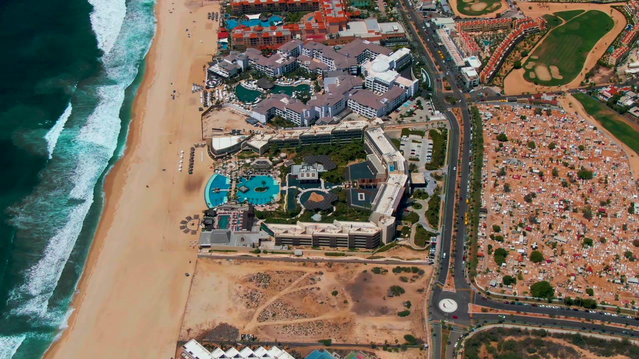 Aerial view over San Jose del Cabo with the beach and waves on the left, resort pools near center, a coastal road and roundabout, sandy lots, and a green landscaped area inland, with no visible people.