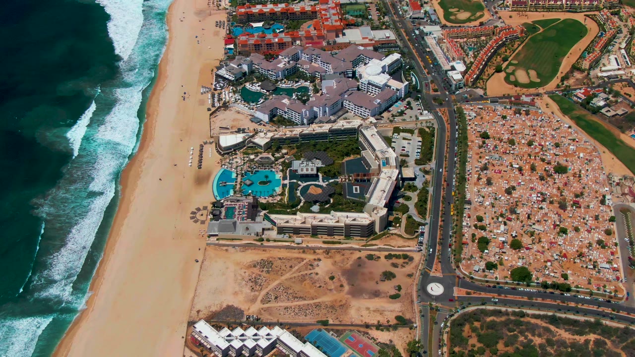 San Jose del Cabo aerial frame showing a darker ocean band, white breaking waves, the sandy beach, resort buildings, road junction, and built-up lots inland, with no visible people.