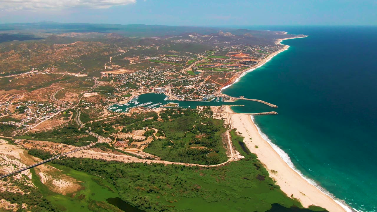 Aerial frame over San Jose Del Cabo with the harbor centered below, beach and surf line along the right edge, inland roads and low buildings beyond, and no visible people.