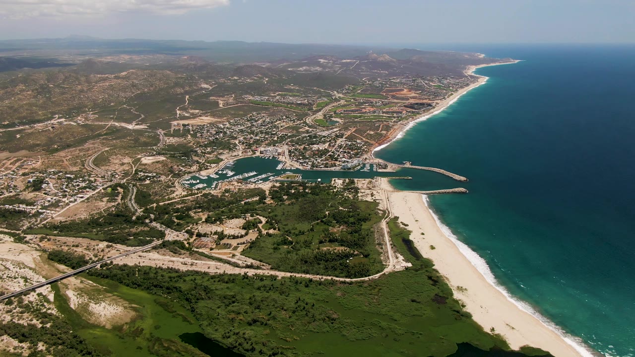 High aerial view over San Jose Del Cabo, Baja California Sur, showing blue ocean, a sandy beach, harbor jetties, roads, green vegetation, dry hills, and no visible people.