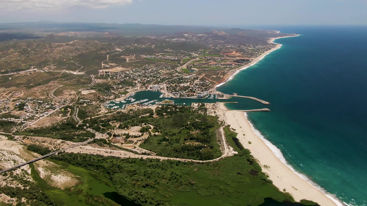 A slightly later aerial frame of San Jose Del Cabo with the harbor centered below, the beach running along the right edge, inland roads and hills beyond, and no visible people.