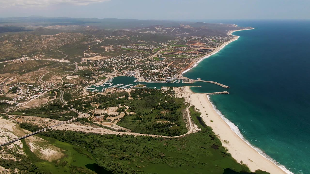 Wide aerial of harbor, beach, ocean, roads, vegetation, and hills.